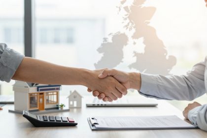 Two people in business attire shake hands over a desk with a contract, calculator, and model houses. A map is visible in the background.