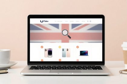 A laptop on a white table displays a website with the Union Jack flag and a magnifying glass icon. Below are images of smartphones and a laptop. A coffee cup, notebook, and smartphone are beside the laptop.