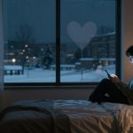 A person sits on a bed using a smartphone in a dimly lit room. Outside the window, it's snowing with a heart shape on the glass.