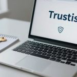 A laptop on a desk displays the word "Trustis" with a shield icon. Next to it, a gold key rests on a lined notepad.