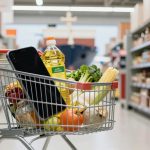 A shopping cart filled with groceries, including a bottle of cooking oil, fresh vegetables, and fruits. A smartphone rests on top. The cart is in a supermarket aisle lined with shelves of various packaged goods.