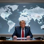 A man in a dark suit and red tie sits at a desk with papers, a phone, and a microphone. Behind him is a large illuminated world map. An American flag is visible to the right.