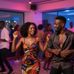 A group of people dancing in a dimly lit club with colorful lights. A woman in a vibrant patterned dress and a man in a dark shirt are smiling and dancing at the center. Other dancers are in the background, with large windows showing city lights outside.