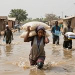 A woman carrying sacks on her head walks through a flooded street, followed by four men holding bottled water and supplies. Muddy water surrounds them, and buildings line the street.