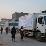 A street scene at dusk with people walking near two white trucks covered with fabric. A woman carries a child in her arms. In the background, a fire burns and more people walk. Buildings line the street, and debris is visible on the ground.