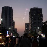 A busy street scene at dusk with people gathered around a food stall illuminated by bright lights. Tall buildings tower on either side, and fireworks are visible in the sky. Colorful shop signs line the street, and cars are parked along the road.