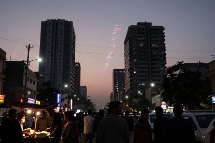A busy street scene at dusk with people gathered around a food stall illuminated by bright lights. Tall buildings tower on either side, and fireworks are visible in the sky. Colorful shop signs line the street, and cars are parked along the road.