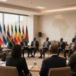 A group of men in suits sit in a semi-circle during a meeting, with multiple African and regional flags displayed behind them. A map of Africa is visible on the wall.