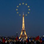 The Eiffel Tower illuminated at night, with a circle of yellow stars above it. People in the foreground hold red and blue flags.