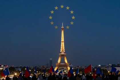 The Eiffel Tower illuminated at night, with a circle of yellow stars above it. People in the foreground hold red and blue flags.