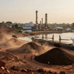 A docked cargo ship is being loaded with piles of red soil at an industrial port. In the background, there are large industrial structures and buildings. Heavy machinery and workers are visible near the soil piles, with a dusty atmosphere suggesting active loading operations. The scene is set beside a body of water under a clear sky.