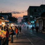 Street market at dusk with vendors and customers at stalls under blue tents, selling colorful fruits. Buildings and graffiti-lined walls in the background.
