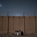 Four people sit on the ground at night, facing a tall concrete wall topped with barbed wire. Two drones with lights fly overhead.