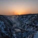 Snow-covered quarry with multiple tunnel entrances carved into rocky cliffs. The sun sets on the horizon, casting an orange glow.