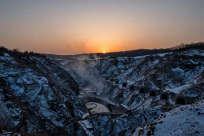 Snow-covered quarry with multiple tunnel entrances carved into rocky cliffs. The sun sets on the horizon, casting an orange glow.