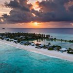 Aerial view of a tropical island at sunset, with sandy beaches, palm trees, and small houses. The turquoise ocean surrounds the island, and dark clouds hover above with the sun setting in the background.