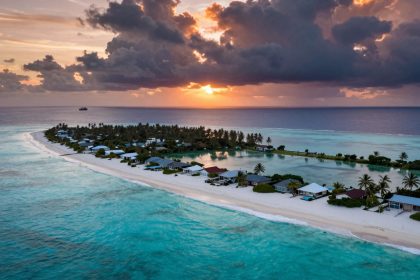 Aerial view of a tropical island at sunset, with sandy beaches, palm trees, and small houses. The turquoise ocean surrounds the island, and dark clouds hover above with the sun setting in the background.