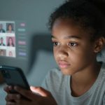 A young girl with curly hair is focused on a smartphone. A holographic display shows a grid of blurred profile pictures in the background.
