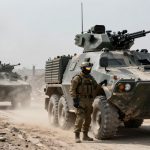Military vehicles in a dusty, urban setting, led by a soldier in tactical gear and helmet. The vehicles are armored and camouflaged.
