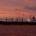 A large cargo ship sails on the ocean under a pink and orange sunset sky. Three drones fly above, and a faint contrail is visible in the sky.