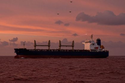 A large cargo ship sails on the ocean under a pink and orange sunset sky. Three drones fly above, and a faint contrail is visible in the sky.