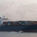 A large cargo ship carrying stacked shipping containers sails on the water. A small boat travels alongside. A faint world map overlays the sky.