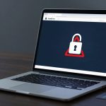 A laptop on a wooden table displays a screen with a padlock icon inside a red triangle, indicating security. A credit card is beside it.