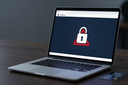 A laptop on a wooden table displays a screen with a padlock icon inside a red triangle, indicating security. A credit card is beside it.
