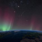 Earth's horizon with vivid green and red auroras in the atmosphere, starry night sky above with visible constellations and stars.