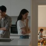 A man and woman stand in a kitchen looking concerned, with arms crossed and hands on chins. In the background, two children sit on a bed playing with toys.