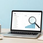 A laptop on a desk displays a bar graph with a magnifying glass overlay. Nearby are a white mug and a spiral notebook. The background is light blue.