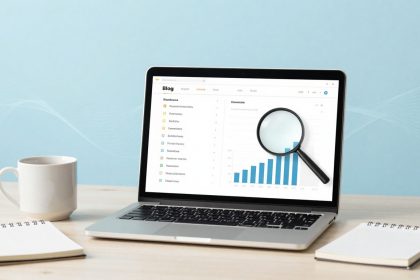 A laptop on a desk displays a bar graph with a magnifying glass overlay. Nearby are a white mug and a spiral notebook. The background is light blue.