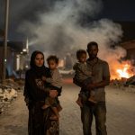 A family of four walks through a rubble-filled street at night. The mother and father each carry a child. Smoke and fire are visible in the background, with damaged buildings on either side.