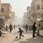 A bustling street scene features a child playing soccer in the foreground. People walk along the dusty road lined with old stone buildings. On the right, a bread vendor sells loaves under a canopy. The atmosphere is hazy, suggesting a busy market setting.
