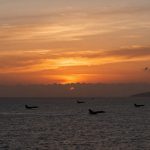 Jets fly over the ocean at sunset, with an orange and yellow sky. Dark clouds and a distant landmass are visible on the horizon.