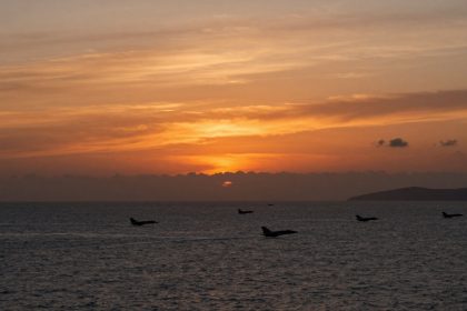 Jets fly over the ocean at sunset, with an orange and yellow sky. Dark clouds and a distant landmass are visible on the horizon.