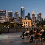 A group of people sitting on chairs outdoors, facing a projector screen on a brick wall. In the background, a city skyline is visible at dusk, with illuminated skyscrapers and a landmark resembling the Brandenburg Gate. The setting suggests a rooftop or open-air venue.
