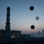 Industrial landscape at dusk with two tall chimneys and a building emitting light. Three floating spheres are in the sky, and silhouettes of people holding flags are visible in the foreground.
