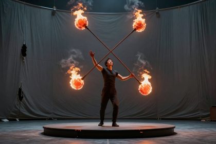 A performer balances a large cross-shaped fire staff with four flaming ends on a circular stage. The background is a dark draped curtain.