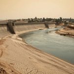 A concrete dam with a curved river channel, surrounded by dry, cracked earth. Buildings and trees are visible in the background under a hazy sky.