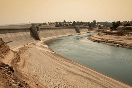 A concrete dam with a curved river channel, surrounded by dry, cracked earth. Buildings and trees are visible in the background under a hazy sky.