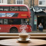 A steaming cup of coffee sits on a wooden table by a window. Outside, a red double-decker bus passes by on a rainy day, and a person is seated across the street, talking on a phone.