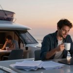 A man sitting on a yacht at sunset, holding a mug and smiling at a laptop. Papers are scattered on the table. Another person is visible inside, working at a desk.