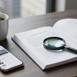A steaming cup of coffee sits on a wooden table next to an open book with a magnifying glass on it. A smartphone displaying a news app is also on the table.