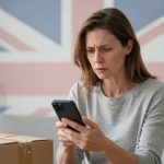 A woman in a grey sweater looks intently at her smartphone. A cardboard box is in the foreground. A Union Jack flag is in the background.