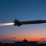 A missile with a bright exhaust trail flies above a silhouetted aircraft carrier and radar dome at sunset, against a twilight sky.