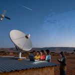 A group of children are gathered around a laptop on a corrugated metal roof under a large satellite dish. A satellite is visible in the sky, with a faint world map overlay. One child is holding a phone. The setting is a desert landscape with trees and mountains in the background, under a twilight sky.