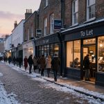 Cobblestone street at dusk with snow on the ground, people walking, and a lit café visible through windows. Signs read "To Let."