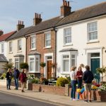Terraced houses with brick and cream facades line a street. Two groups of people stand talking on the sidewalk. Gardens have potted plants and toys.