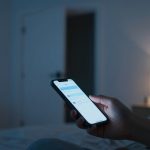 A person holds a smartphone in a dimly lit bedroom. The phone screen displays a messaging app. A blurred lamp glows softly in the background.